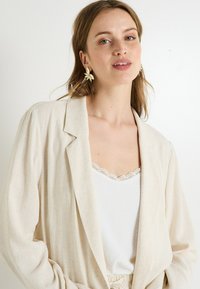 Blazer de lino beige con un corte relajado y solapas con muescas, usado sobre una camiseta de tirantes blanca con encaje. Pendientes de hojas doradas acentúan el look.