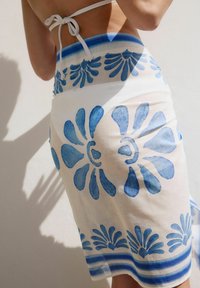 A person’s back is visible, wearing a white bikini top and a skirt featuring large blue floral patterns against a light background.