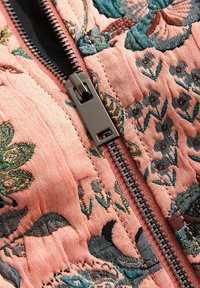 Gewatteerde stof in lichtroze met ingewikkeld geborduurde bloemen. Voorzien van een metalen rits met een donkere trekker en een contrasterende voering.