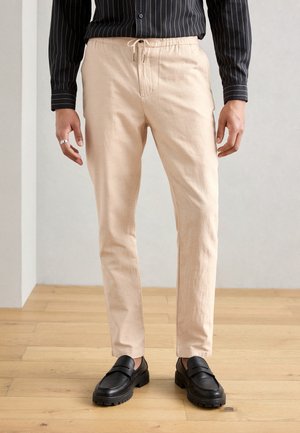 Homme portant un pantalon beige à cordon, chemise noire rayée, mocassins en cuir noirs, debout sur un sol en bois clair contre un mur uni.