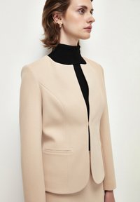 Blazer beige su misura con scollatura rotonda, due tasche frontali e tessuto testurizzato, indossato sopra un dolcevita nero.