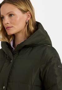 Olīvu zaļš pūkains jaka ar augstu apkakli un kapuci, ar sudraba spiedpogām un tekstūru vīlēm. Logo detaļa uz piedurknes.