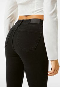 Jeans negros de cintura alta con una silueta ajustada, que cuentan con bolsillos traseros y una etiqueta texturizada en la cinturilla. Acabado de tela suave.