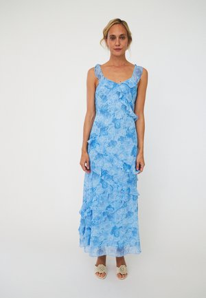 Femme porte une robe bleu fleurie sans manches avec des volants et des sandales blanches avec détail floral, debout devant un fond blanc uni.