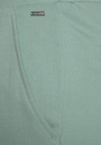 Svetlozelená tkanina s hladkou textúrou, obsahujúca trojuholníkovú kapsu a malý kovový logo akcent. Detailne viditeľné švy.