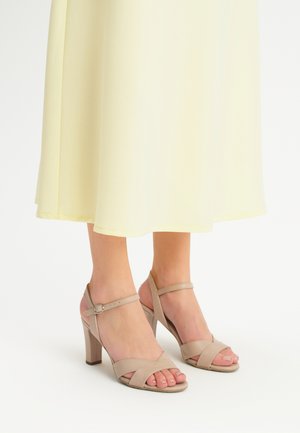 Sandalias de tacón alto beige con tiras cruzadas, hebilla en el tobillo y textura suave, combinadas con un vestido amarillo fluido.