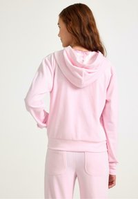 Różowy hoodie wykonany z miękkiego materiału, wyposażony w kaptur ze sznurkiem i ściągaczami na rękawach. Tył jest gładki, a krój luźny, uzupełniony pasującymi spodniami.