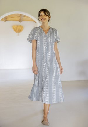 Femme souriante marchant à l'intérieur, portant une robe midi à manches courtes, boutonnée et à motif, avec des ballerines beiges.