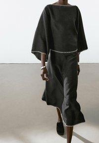 Tenue deux pièces noire, ample, avec des manches larges et un ourlet courbé; présente des coutures blanches contrastantes le long des bords.