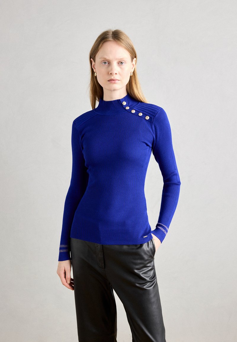 Ribbad långärmad topp i klart blått, med hög krage, dekorativa knappar på axeln och kontrasterande detaljer vid ärmsluten.