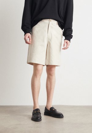 Beżowe szorty do kolan wykonane z tkaniny o fakturze, w zestawieniu z czarnymi loafersami ozdobionymi złotymi detalami. Model stoi na neutralnym tle.