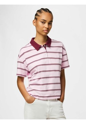 Jeune personne aux cheveux tressés portant un polo rayé rose et bordeaux et un pantalon blanc, debout avec les mains dans les poches.