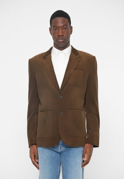 Blazer en velours côtelé marron avec un col à revers, deux poches avant et une fermeture à un bouton. Porté sur une chemise blanche à col et un jean bleu.