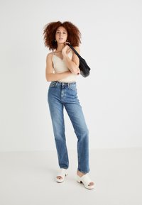 Lichtbeige kanten tanktop, hoog getailleerde blauwe spijkerbroek en witte block-heeled muilen met een open teen. Zwarte handtas.