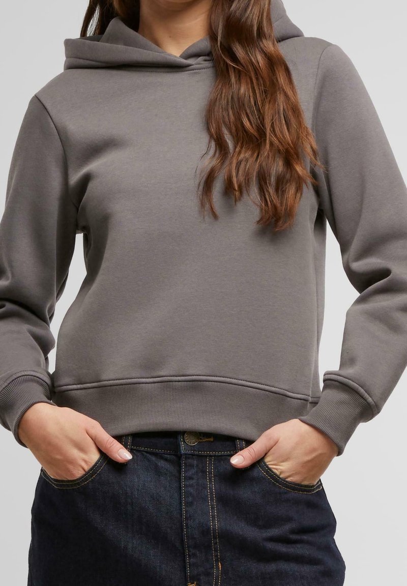 Personne portant un sweat à capuche gris avec les mains dans les poches d'un jean bleu foncé, montrant les détails du haut du corps et du milieu.