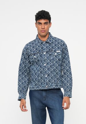 Giochino in denim con un motivo geometrico bianco, colletto classico, tasche frontali e bottoni argentati. Abbinato a jeans blu scuri.