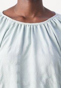 Blouse bleu clair en tissu texturé, avec un col froncé et une belle chute. Présente des motifs subtils et une coupe décontractée et fluide.