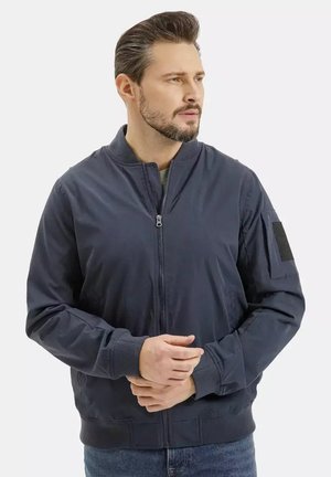 Mężczyzna z brodą i wystylizowanymi włosami, ubrany w granatową kurtkę bomberkę na dżinsach, patrzący w prawo, z rękami złączonymi przed sobą.