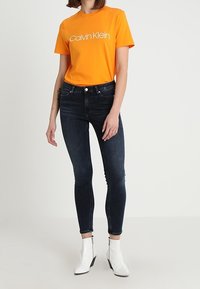 Camiseta de algodón naranja con el logo blanco de "Calvin Klein", combinada con jeans skinnies oscuros. Botines blancos con cremalleras.
