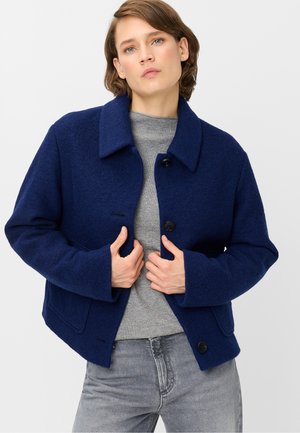 Woman with short brown hair wearing a navy blue buttoned wool jacket over a gray sweater and gray jeans against a plain light background.