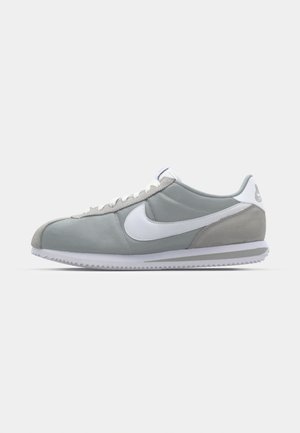 Chaussure de sport grise avec une tige en daim et textile, logo Nike en blanc et lacets blancs. Elle dispose d'une semelle en caoutchouc texturée et d'un collier rembourré.