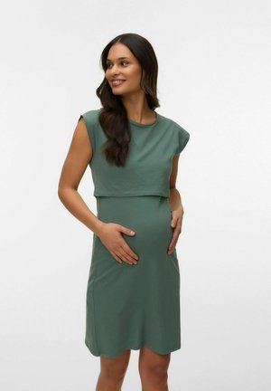 Gravid kvinde i grønnærmeløs kjole, der blidt holder maven, smiler og kigger til siden mod en ensfarvet baggrund.