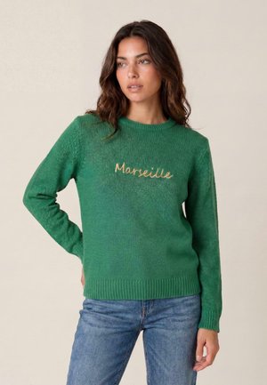 Donna con capelli castani ondulati che indossa un maglione verde in maglia con "Marseille" ricamato in beige, abbinato a jeans blu, in piedi davanti a uno sfondo semplice.