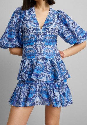 Femme portant une robe bleue à motifs floraux avec manches bouffantes, encolure en V, taille ajustée et jupe à volants à étages sur un fond gris uni.