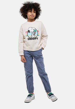 Enfant aux cheveux bouclés debout, main dans la poche, portant un sweat-shirt Snoopy crème, un pantalon de jogging bleu et des baskets blanches à lanières vertes.