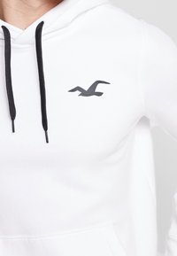 Vit hoodie med svarta snören och en liten svart logga på bröstet. Tyget verkar slätt och mjukt, lämpligt för avslappnad användning.