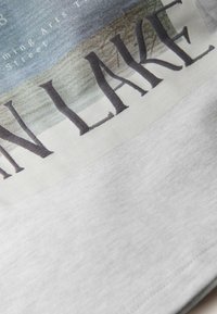 Graues Baumwoll-T-Shirt mit einem malerischen Druck eines Sees, auf dem am unteren Rand in fettem Schwarz der Text "SEE" deutlich sichtbar ist.