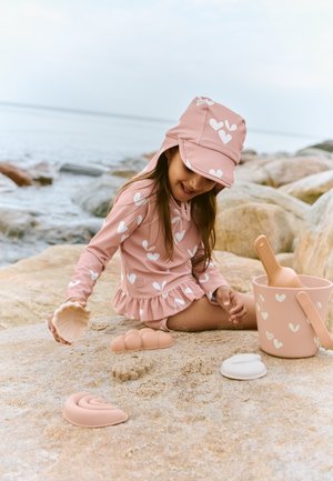 Rožinė plaukimo marškinėliai su širdelių raštais ir raukinėta apatine dalimi, derantis kibirėlis su rankena ir smėlio žaislai, įskaitant kriauklių ir bangų formų, ant smėlėtos paplūdimio.