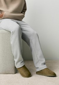 Grå sweatpants med afslappet pasform, parret med brune ruskinds slippers. Figuren sidder på en lys, struktureret skammel. Blød, neutral baggrund.