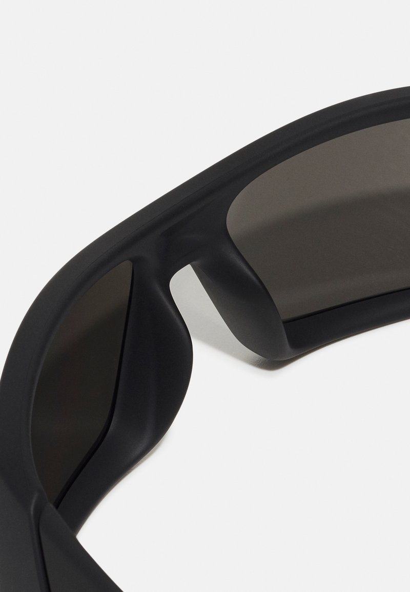 Gafas de sol negras con un acabado mate; montura ancha con bordes curvados y lentes oscuros, diseñadas con contornos elegantes y suaves.