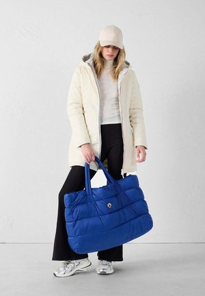 Borsa a spalla blu imbottita con due manici robusti, con una texture trapuntata. Il modello indossa un cappotto crema, pantaloni neri e sneaker argentate.