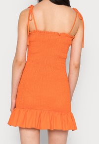 Společenské mini šaty v oranžové barvě s vypasovaným střihem, nastavovacími šňůrkami na ramínkách a řaseným lemem. Tkanina má strukturovaný povrch.