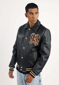 Veste en cuir noir avec un col et des poignets côtelés, ornée d'accents brodés et de détails dorés. Fermeture à boutons sur le devant avec poches latérales.