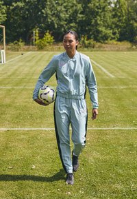 Costum sportiv de culoare albastru deschis, cu dungi negre pe lateral, care include o jachetă cu fermoar și pantaloni cu talie elastică, ținând în mână o minge de fotbal cu model.