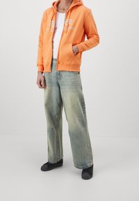 Felpa arancione con zip, tasche frontali e grafiche bianche, abbinata a jeans in denim blu chiaro larghi e calzature nere.