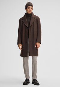 Bruine wollen jas met grote kraag, drie knopen en zijzakken, gedragen over een donkere turtleneck en lichtgrijze geklede broek met een patroon. Zwarte leren schoenen.