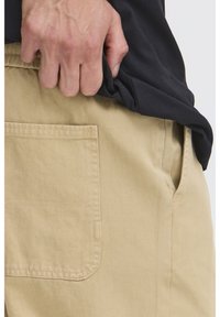 Pantaloni di cotone beige con tasca laterale e cuciture visibili. Parzialmente visibile una mano che afferra l'orlo di una camicia nera.
