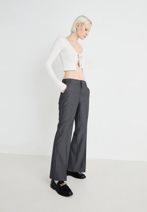 Femme aux cheveux blonds courts portant un top court blanc à manches longues et un pantalon large gris à fines rayures avec des mocassins noirs.