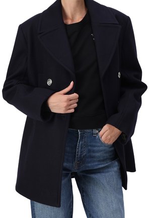 Person iført en mørk marineblå dobbeltradet frakke over en sort skjorte og blå jeans, med den ene hånd, der justerer frakken og den anden i en lomme.