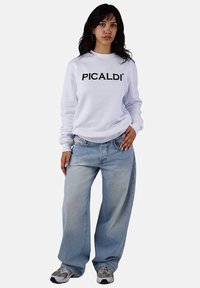 White sweatshirt with black "PICALDI" logo, paired with light blue wide-leg jeans and gray sneakers. Model standing with hand on hip.