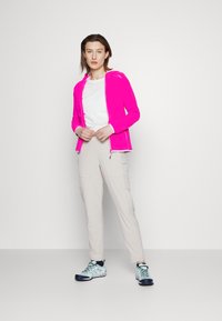 Giacca zip con cappuccio di un vivace colore rosa, indossata sopra una maglietta bianca. Pantaloni cargo beige e scarpe da ginnastica multicolori completano l'outfit.