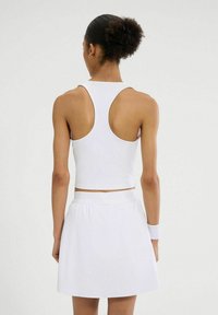 Biały sportowy crop top z okrągłym dekoltem i designem racerback, w zestawie z dopasowaną białą spódnicą. Gładka tekstura i dopasowany krój.