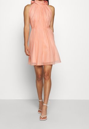 Kvinde iført ærmeløs ferskenfarvet halterkjole med plisseringer, kombineret med hvide høje hæle sandaler med stropper, står foran en ensfarvet baggrund.