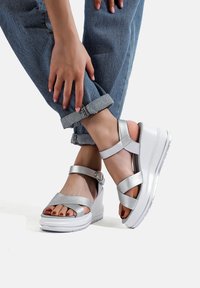 Sandali platform argento con una finitura liscia, fasce incrociate, una fibbia regolabile alla caviglia e una suola bianca e testurizzata. Jeans a maniche risvoltate mostrati.