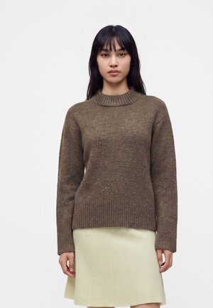 Femme aux longs cheveux foncés portant un pull en maille marron et une jupe côtelée écrue, debout devant un fond clair uni.