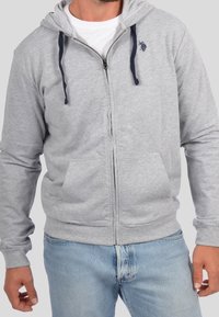 Sudadera con cremallera gris, con cordones azul marino, bolsillos frontales y un pequeño logo en el pecho. La tela parece suave y texturizada.